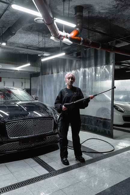 A woman uses a pressure washer on luxury cars in an indoor car wash setting.